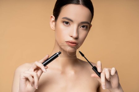 Confident asian woman with naked shoulders holding mascara and applicator isolated on beige