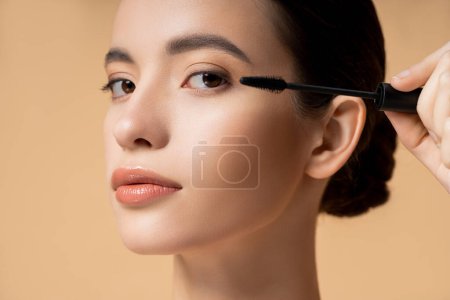 Close up view of young asian woman applying mascara and looking at camera isolated on beige