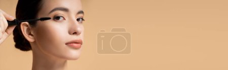 Jolie femme asiatique appliquant mascara et regardant la caméra isolée sur beige, bannière