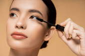 Young asian model with natural makeup and perfect skin holding mascara applicator isolated on beige Poster #666744668