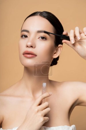 Young asian woman in top applying eyebrow gel and looking at camera isolated on beige