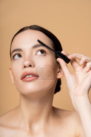 Pretty young asian woman with naked shoulders looking up and apply eyebrow gel isolated on beige