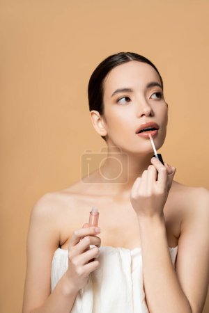 Young brunette asian woman in top applying lip gloss while posing isolated on beige