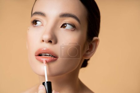 Foto de Retrato de joven modelo asiático aplicando brillo de labios y mirando hacia otro lado aislado en beige - Imagen libre de derechos