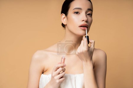 Pretty young asian woman in top looking at camera while applying lip gloss isolated on beige