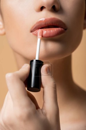 Foto de Vista recortada de la mujer joven que aplica brillo de labios mientras hace el maquillaje aislado en beige - Imagen libre de derechos