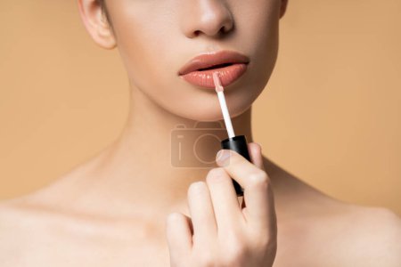 Cropped view of young model with naked shoulders applying lip gloss isolated on beige