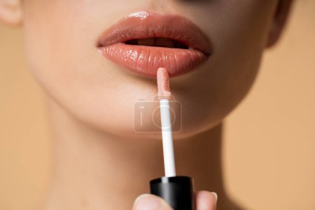 Close up view of young woman applying lip gloss while doing makeup isolated on beige