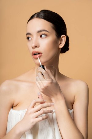 Pretty asian model with naked shoulders in top applying lip gloss and looking away isolated on beige
