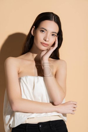 Young brunette woman in top looking at camera while posing on beige background with shadow