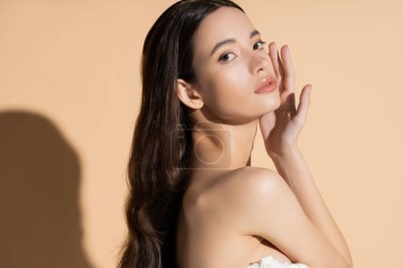 Pretty asian woman with long hair and naked shoulder touching cheek and standing on beige background