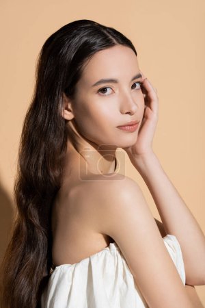 Portrait of young asian woman in top looking at camera and touching face on beige background