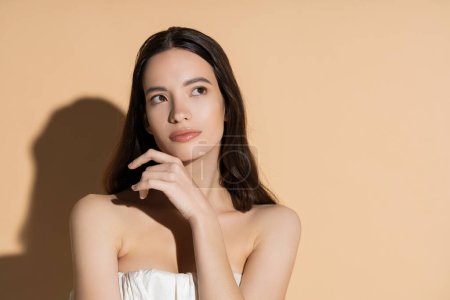 Junge langhaarige asiatische Frau mit natürlichem Make-up posiert auf beigem Hintergrund mit Schatten