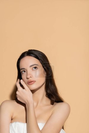 Young asian woman with long hair and natural makeup touching cheek on beige background with shadow