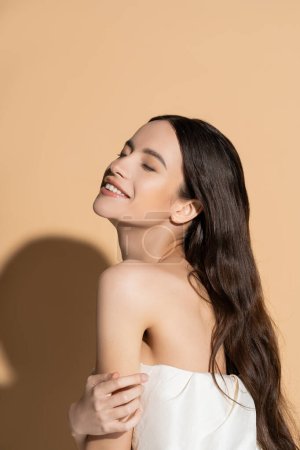 Sonriente y de pelo largo mujer asiática con hombro desnudo de pie sobre fondo beige con sombra