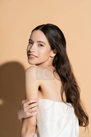 Positive young asian woman touching shoulder and looking at camera on beige background with shadow