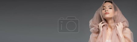 Élégante jeune femme asiatique regardant la caméra et posant avec un tissu beige isolé sur gris, bannière