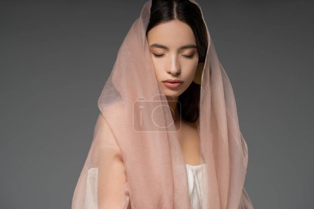 Portrait of pretty young asian woman with closed eyes posing with beige cloth isolated on grey