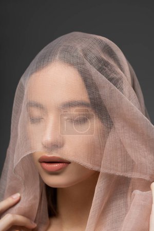 Portrait of young and beautiful asian woman with beige cloth on head standing isolated on grey