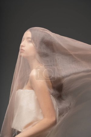Pretty young asian woman in top posing with beige fabric on head while standing isolated on grey