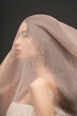 Brunette asian model in top posing under beige fabric on head isolated on grey tote bag #666745610