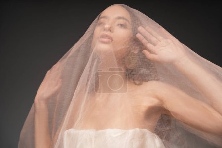 Photo for Asian model with naked shoulders posing under beige fabric while standing isolated on grey - Royalty Free Image