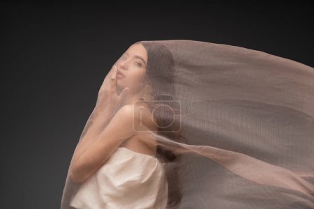 Brunette asian model touching chin and posing under beige cloth isolated on black tote bag #666745698