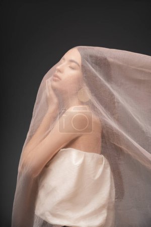 Young asian woman in top touching cheek while posing under beige cloth isolated on grey Mouse Pad 666745778