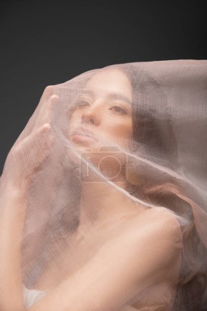 Retrato de joven morena mujer asiática tocando tela beige mientras posando aislado en negro