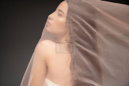 Foto de Vista lateral de modelo asiático con hombro desnudo mirando hacia otro lado mientras posando bajo tela aislada en negro - Imagen libre de derechos