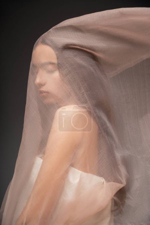 Portrait of pretty asian model with closed eyes posing under beige fabric isolated on black