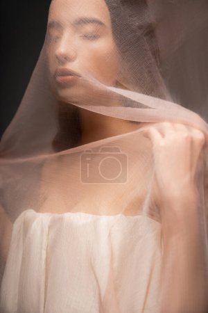Foto de Joven morena modelo asiático con los ojos cerrados vistiendo top y posando bajo tela aislada en negro - Imagen libre de derechos