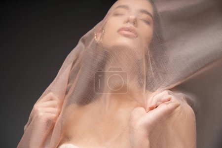 Foto de Retrato de modelo asiático con los ojos cerrados posando bajo tela beige mientras está de pie aislado en negro - Imagen libre de derechos
