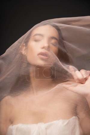 Asian woman with closed eyes and naked shoulders touching beige cloth while posing isolated on black