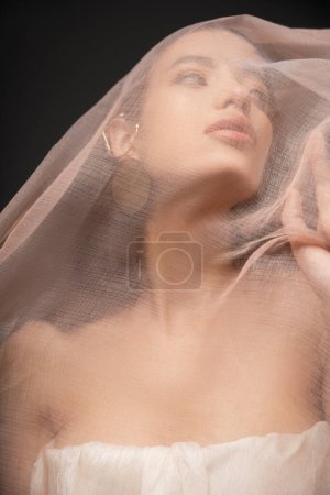 Foto de Vista de ángulo bajo de joven modelo asiático con hombros desnudos mirando hacia otro lado bajo tela aislada en negro - Imagen libre de derechos