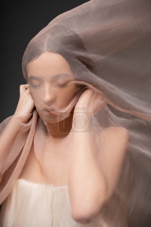 Photo for Portrait of young asian model with closed eyes holding beige fabric and standing isolated on black - Royalty Free Image