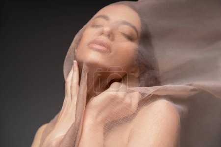 Young asian woman with closed eyes touching neck and posing under beige cloth isolated on black Poster 666745930