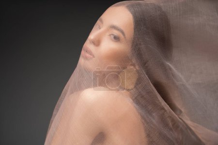 Foto de Joven modelo asiático con hombro desnudo mirando a la cámara bajo tela beige aislado en negro - Imagen libre de derechos