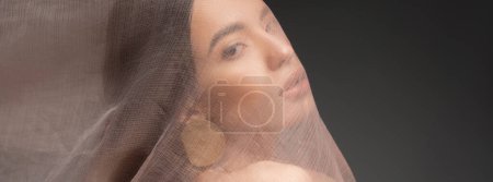 Photo for Asian model with earring looking at camera while posing under cloth isolated on black, banner - Royalty Free Image