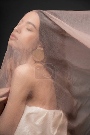 Pretty asian model in top and earring posing under beige cloth while standing isolated on black