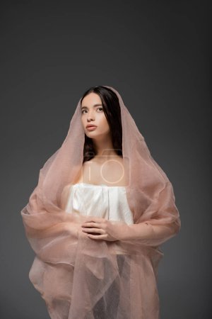 Young brunette asian model in top posing in beige fabric while posing isolated on grey