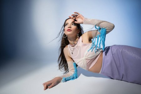 Trendy asian woman with bold makeup and mesh top looking up and lying on blue background