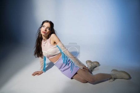 Trendy brunette asian woman in mesh top and skirt looking away while sitting on blue background