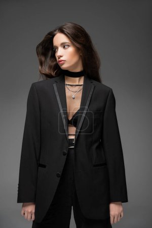 Modelo asiático de moda en top de malla y chaqueta negra mirando hacia otro lado mientras posan aislados en gris