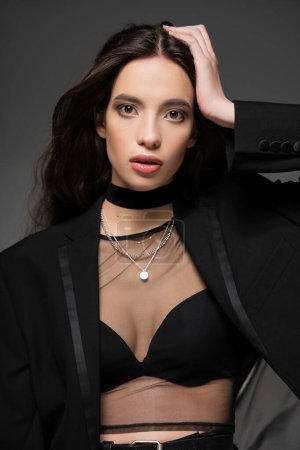 Trendiges junges asiatisches Model posiert in schwarzer Jacke und silbernen Ketten, während es isoliert auf grau steht