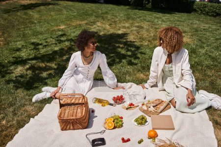positive african american girlfriends near fruits, vegetables on blanket, summer picnic