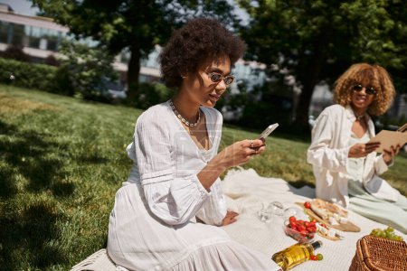 african american woman using smartphone during summer picnic with girlfriend