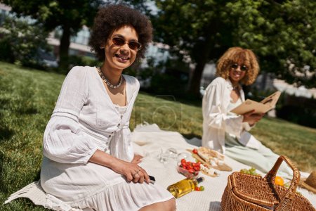 Foto de Mujer afroamericana con teléfono inteligente mirando a la cámara cerca de la novia, picnic de verano - Imagen libre de derechos