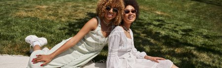 joyful african american girlfriends in sunglasses looking at camera on lawn on summer picnic, banner