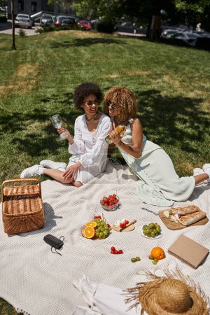 Téléchargez les photos : Copines afro-américaines élégantes sur le pique-nique d'été sur couverture, fruits frais, vin, panier de paille - en image libre de droit
